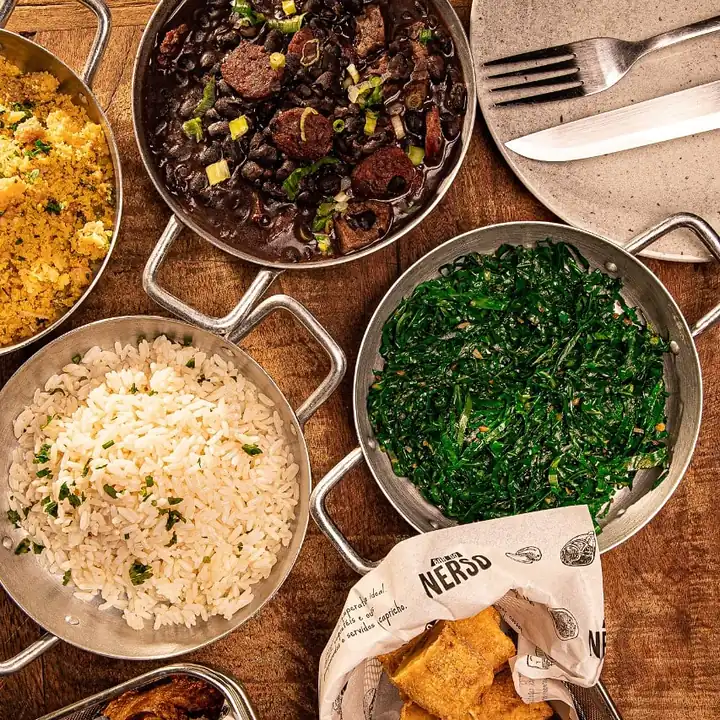 Panelas da casa com feijoada, arroz, farofa e couve servidas no centro da mesa para dividir