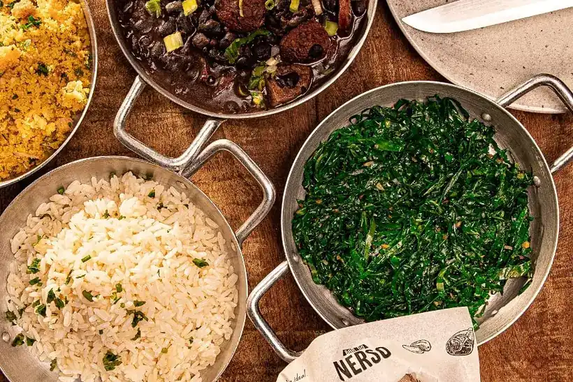 Mesa compartilhada com feijoada, arroz e couve nas panelas da casa